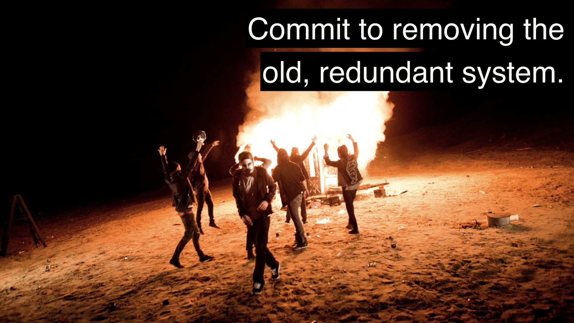 "Commit to removing the old, redundant system."

The photo is of a bunch of people dancing around a bonfire on the beach or desert. Someone in black-and-white clothing and mask is facing the camera.